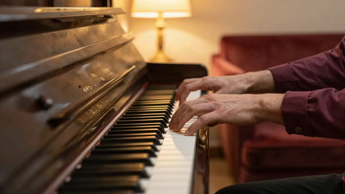 Adult hands playing a chord on a black upright piano in a cozy living room at dusk